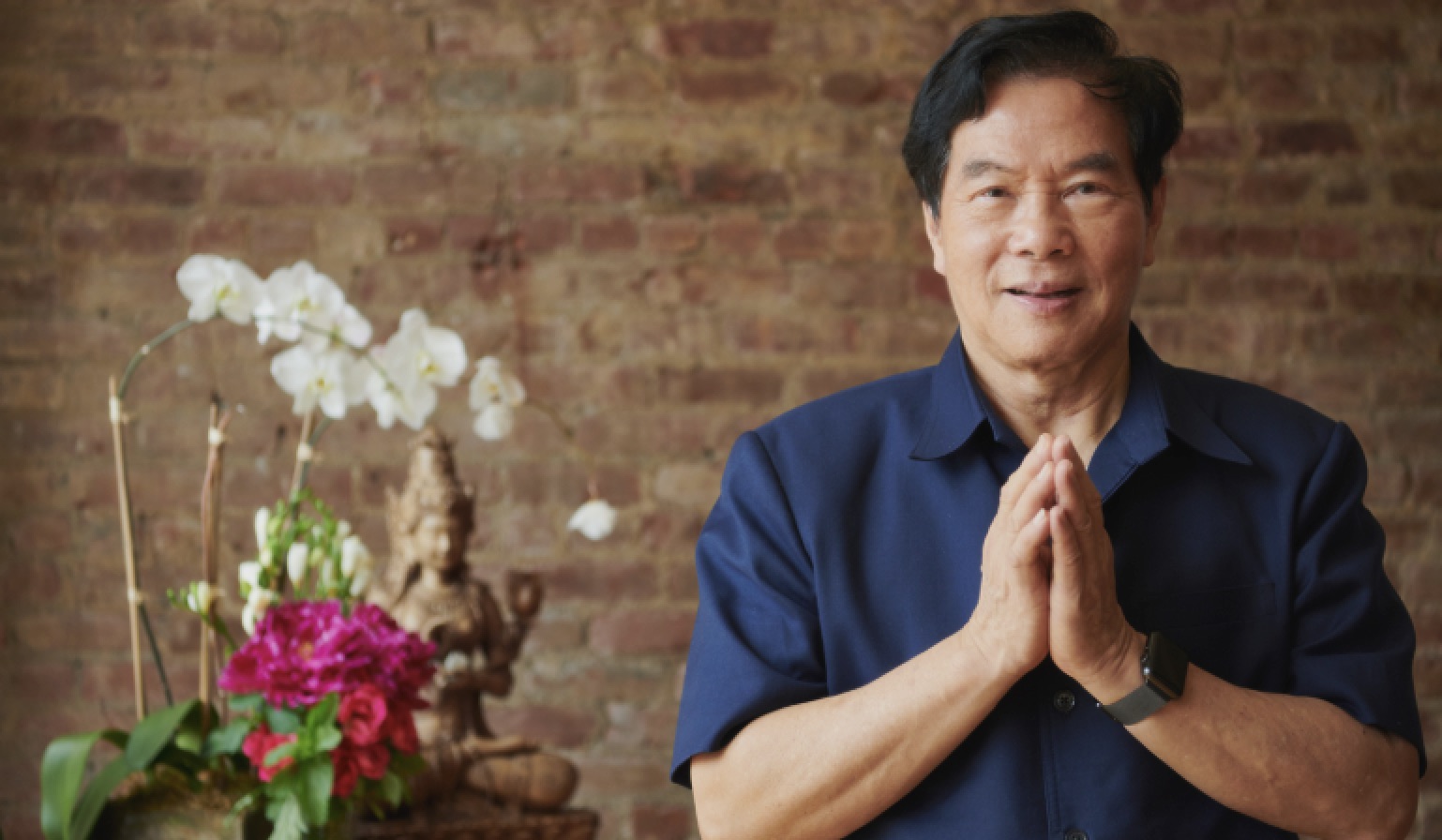 Mantak Chia, hands together, beside orchids and a bronze figure